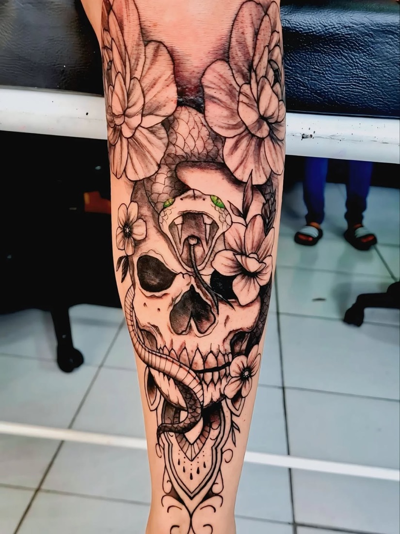 Tatuagem de caveira com flores e cobra em preto e cinza na canela